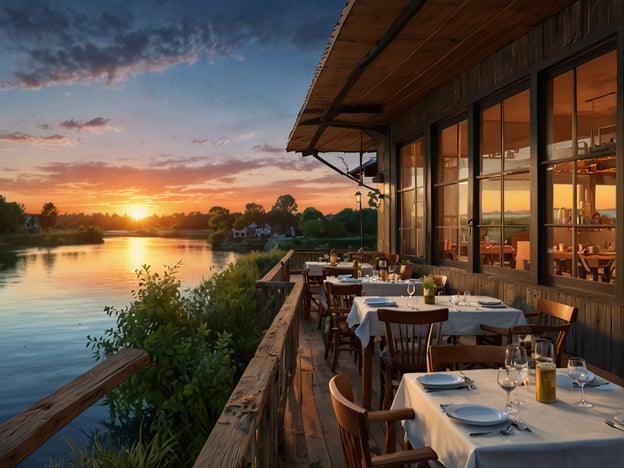 Auf dem Bild ist eine einladende Terrasse eines Restaurants am Flussufer zu sehen. Tische sind liebevoll mit weißen Tischdecken gedeckt und erwarten Gäste, die die kulinarischen Genüsse genießen möchten. Die Atmosphäre wird durch den Sonnenuntergang, der den Himmel in sanften Farbtönen taucht, zusätzlich verschönert. Der Blick auf das ruhige Wasser und die umgebende Natur schafft eine perfekte Kulisse für ein entspanntes Essen im Freien.